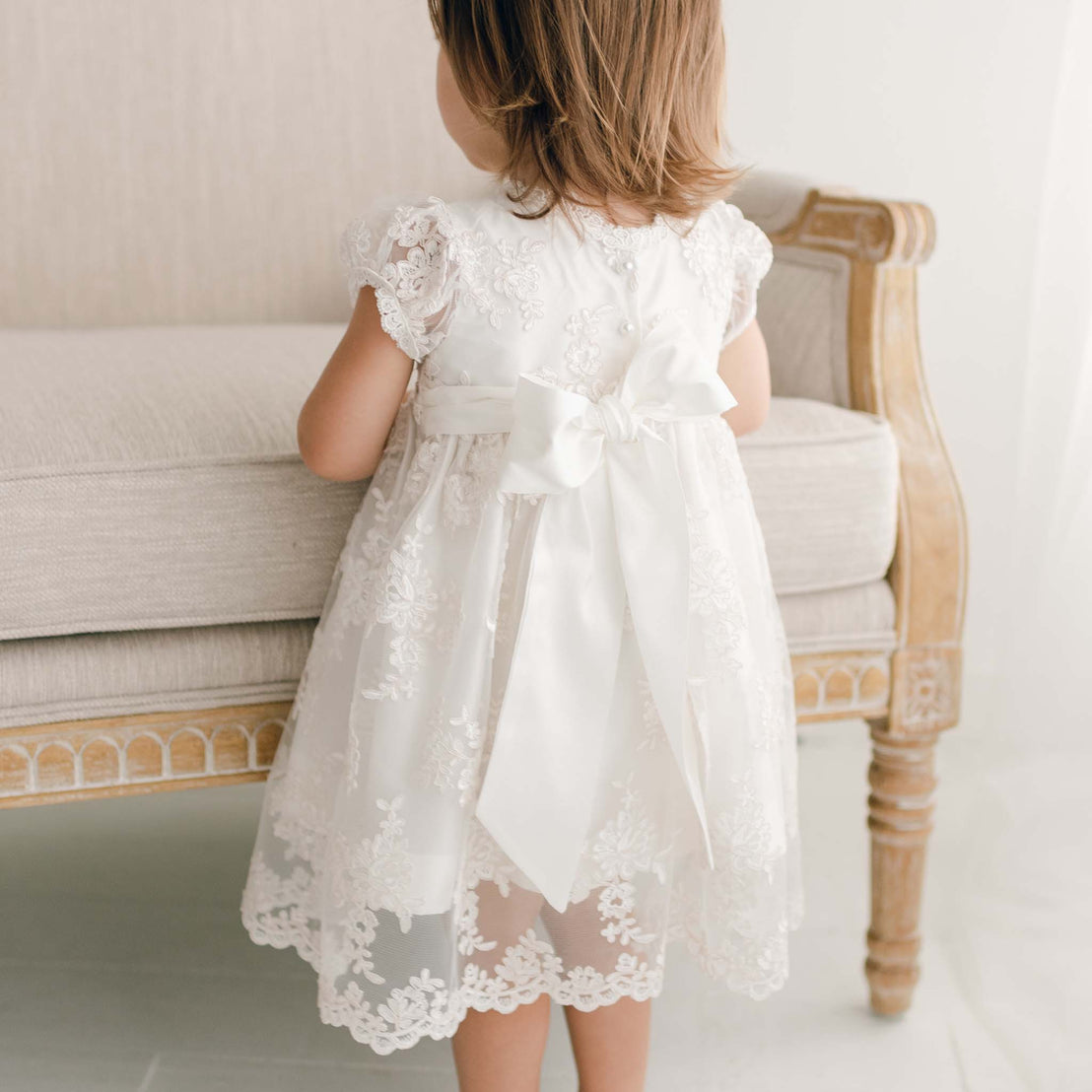 Baby girl sitting by chair wearing Penelope christening dress and bow.