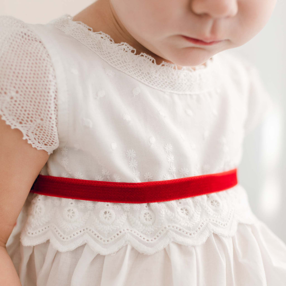 Close-up of a toddler in the Emily Dress & Bloomers with a red sash, focusing on the detailed lace and fabric texture. The child's face is partially visible, looking downward.