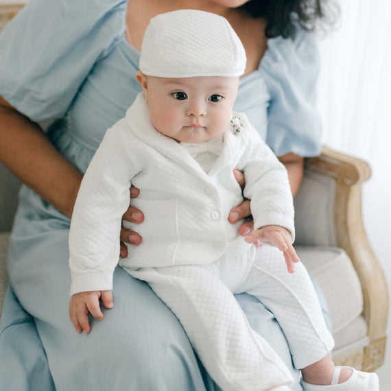 Close up detail of baby boy sitting with his mother and wearing the Elijah 3-Piece Suit made from 100% white textured cotton jacket and pants, and a white cotton / rayon blend shirt. He is also wearing the matching White Velvet Bow Tie & Boutonniere.