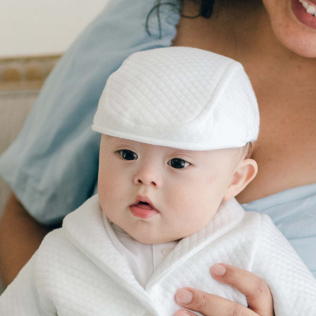 Baby boy sitting on his mother's lap and wearing the Elijah Newsboy Cap. The photo features the top and sides of the hat of the hat in the "newsboy" style in a white textured cotton.
