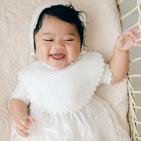 A joyful baby dressed in white, wearing a lace bonnet and handmade Eliza Bib, lies on a textured surface. The baby has a big smile and dark, wavy hair. The outfit is ornate with intricate lace details and floral edge lace, giving a vintage touch and delicate charm.