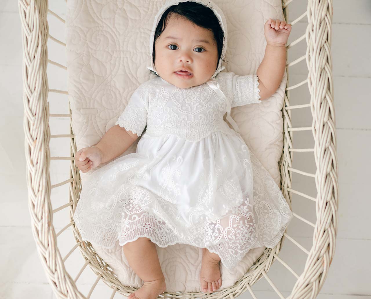 A baby wearing a white lace dress and bonnet lies in a woven basket. The baby looks up with one hand raised, surrounded by a cream-colored quilted cushion.