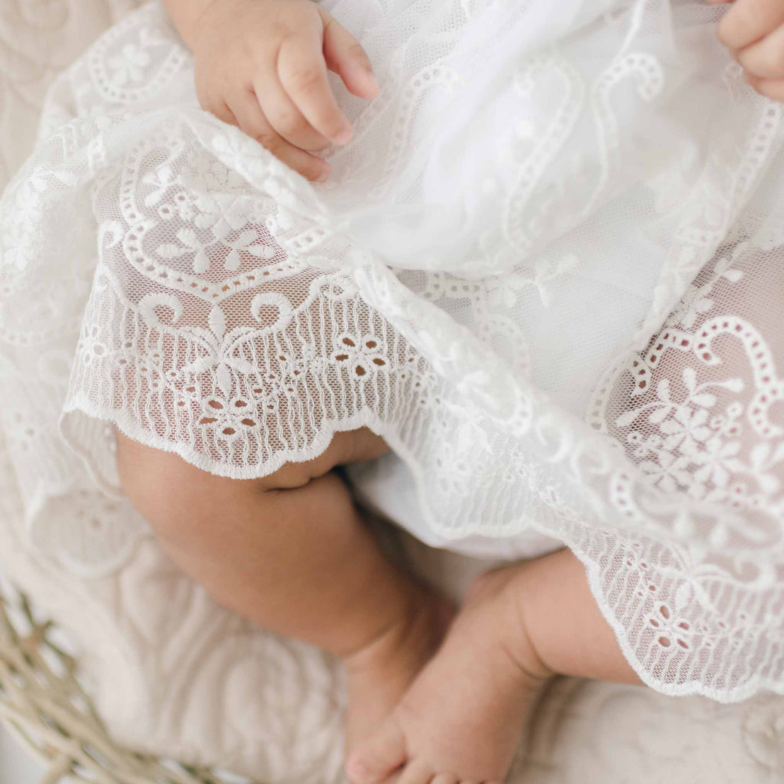 A close-up image of a baby wearing an Eliza Lace Dress & Headband. The baby's small hands are visible gently holding the fabric, and the baby's legs are also partially seen, with tiny feet peeking out from under the dress. The background is soft and neutral.