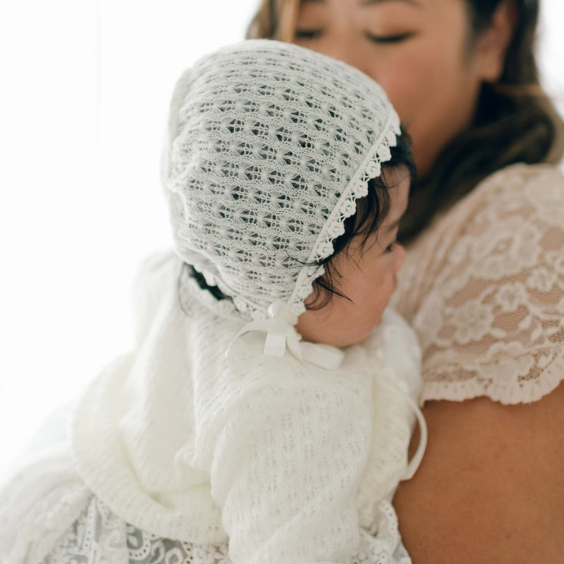 A woman holds a baby who is wearing a white lace bonnet and matching outfit. The baby is facing to the side and looking away from the camera, while the woman, with her face partially visible, lovingly supports the child in an Eliza Knit Sweater. The background is softly lit.