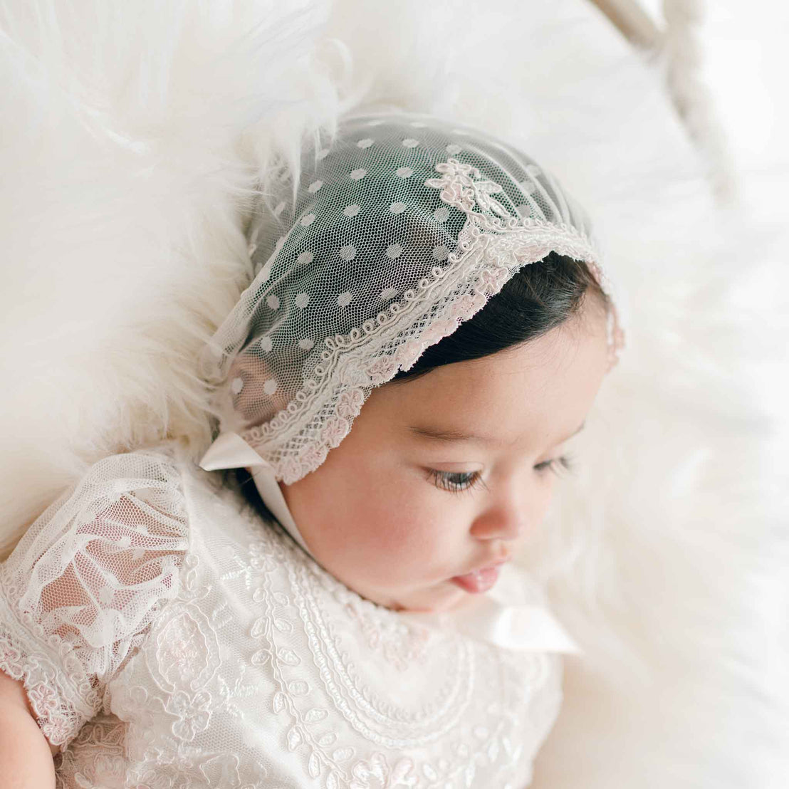 Close-up of a baby wearing a Elizabeth Newborn Christening Bonnet against a soft white background. Details showcase the sheer lace bonnet with silk ribbon ties fit securely around her face, luxurious details characteristic to high end baby clothes.