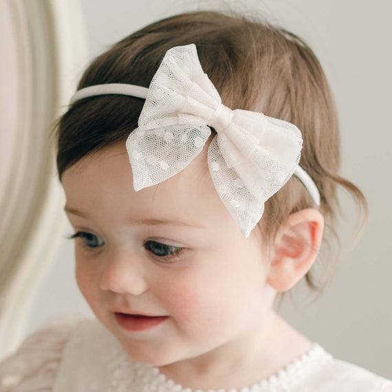 A toddler with brown hair is wearing the Elizabeth Lace Bow Headband, artfully handmade and adorned with delicate embroidered lace. Dressed in a light-colored outfit, the child gazes slightly off to the side with a gentle expression. The blurred, neutral-colored background enhances this tender moment.