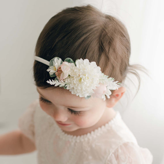 A toddler with short brown hair adorned with an Elizabeth Flower Headband looks down, dressed in a soft, lace-like white dress. The background is softly blurred, emphasizing the child's gentle and delicate appearance.