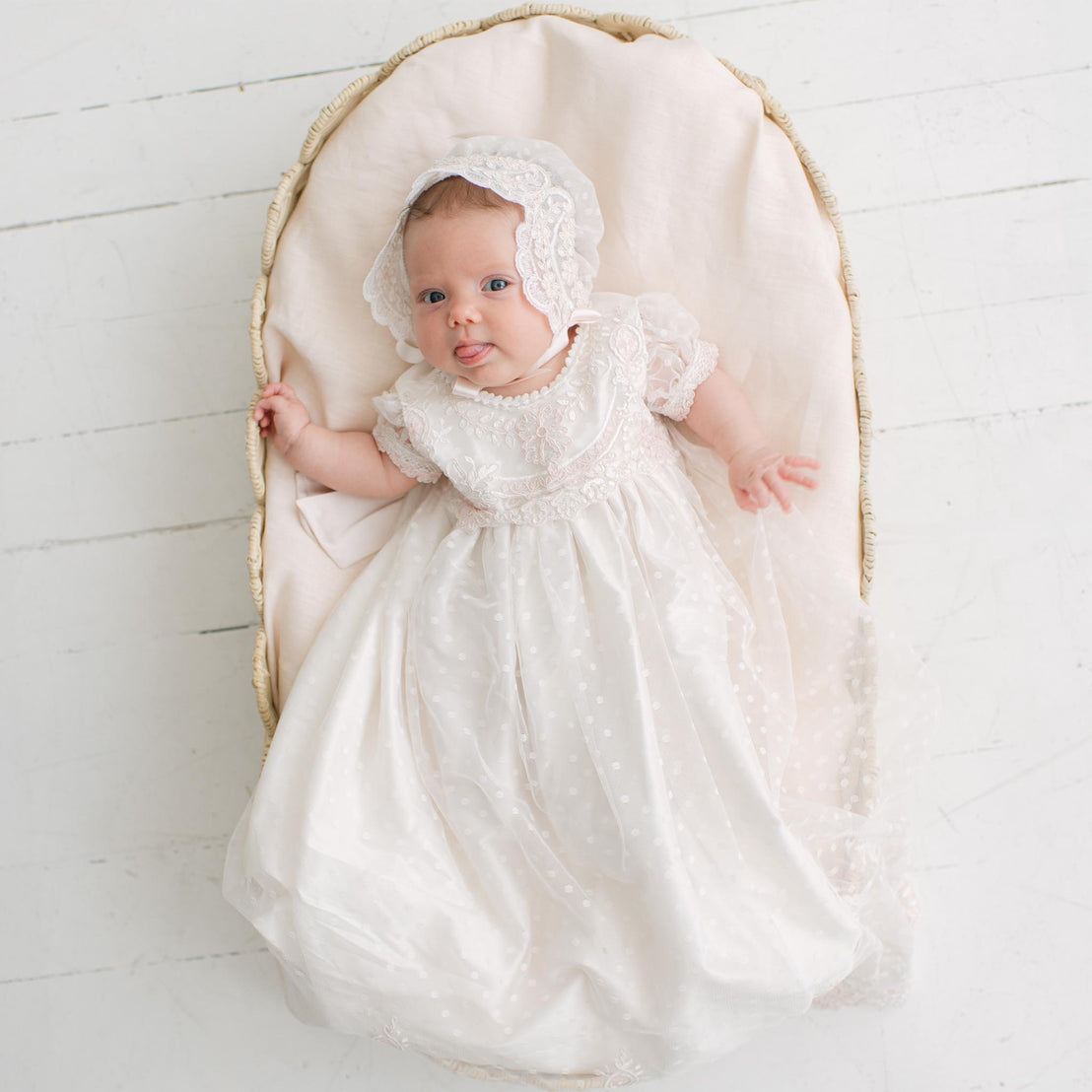 Baby in the Elizabeth Newborn Christening Gown & Bonnet and bonnet lying in a wicker crib on a white wooden floor. Part of the Baby Beau & Belle Baby Girl Christening Dress Collection.
