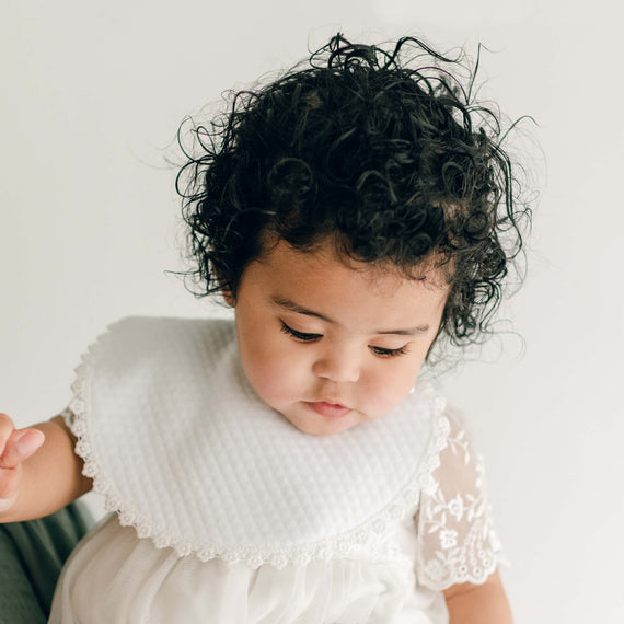 A baby girl with curly dark hair, wearing an Ella Bib and a the Ella Romper Dress, looks down with a thoughtful expression against a plain white background.