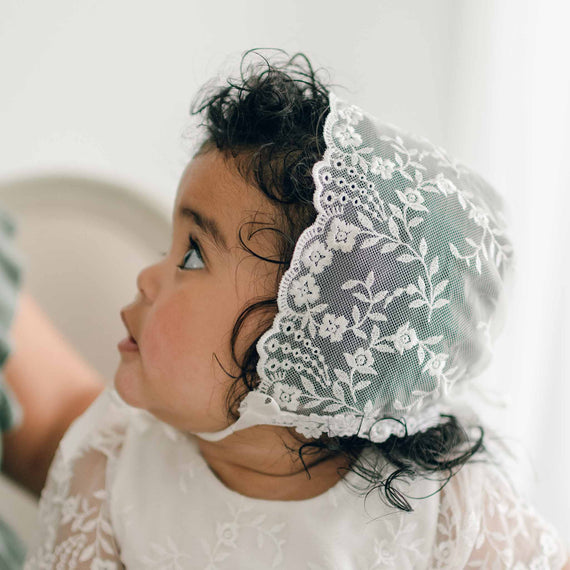 A baby with curly hair wears an Ella Lace Bonnet, adorned with floral embroidered netting, and the matching Ella Christening Gown. The baby looks to the side, revealing the intricate floral pattern on the bonnet. The background is softly blurred, bringing focus to the baby's profile.