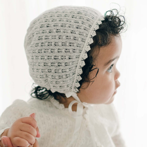 A baby wearing an Ella Knit Bonnet with a ribbon closure, looking to the side. The child has curly dark hair peeking out from under the cotton lace bonnet and is wearing the Ella Knit Sweater, holding onto an adult's hand.
