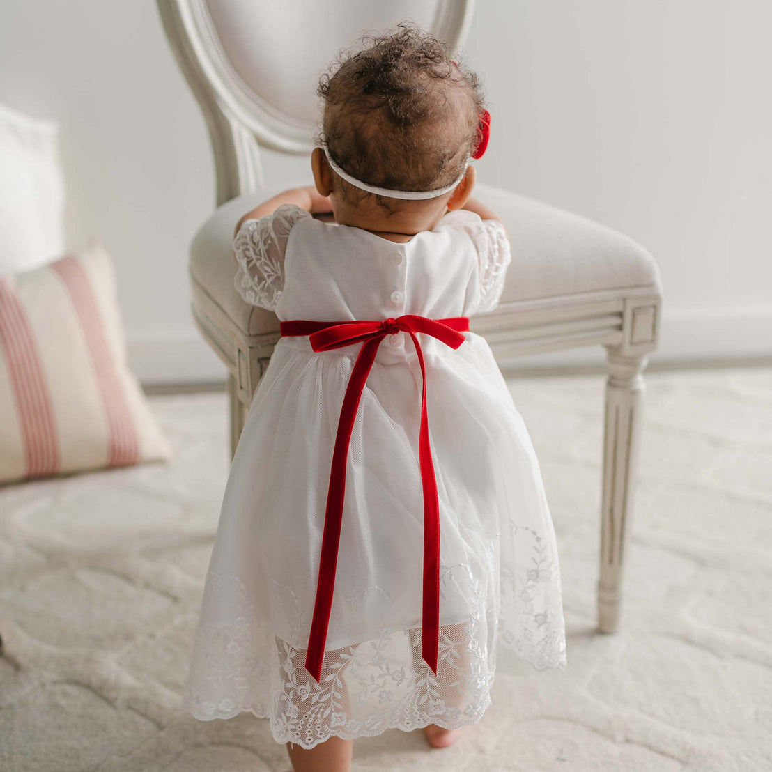 A baby in an Ella Holiday Romper Dress with floral embroidered lace leans against a white chair. The baby's curly hair is adorned with a headband, and the outfit is complemented by a charming red velvet ribbon sash. In the background, a cushion with red stripes sits on a light-colored carpet.