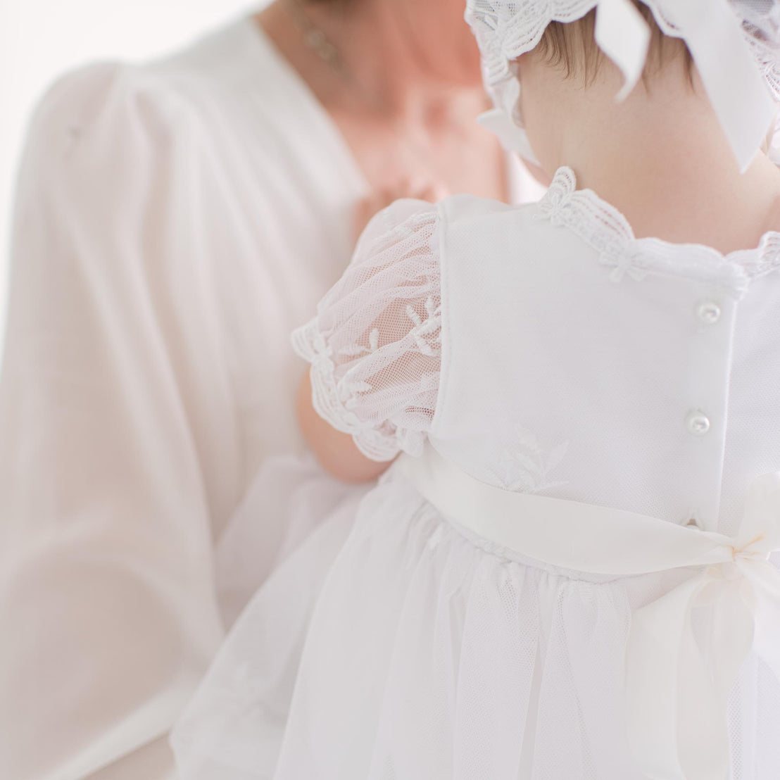 Close-up of a baby in a white dress with lace and pearl button details, held by an adult.