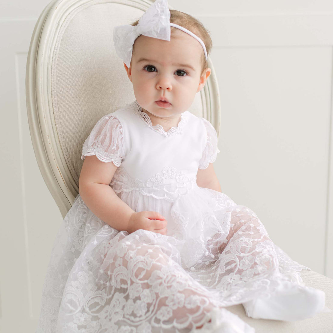 Baby in a white lace dress with a bow headband against a light background.