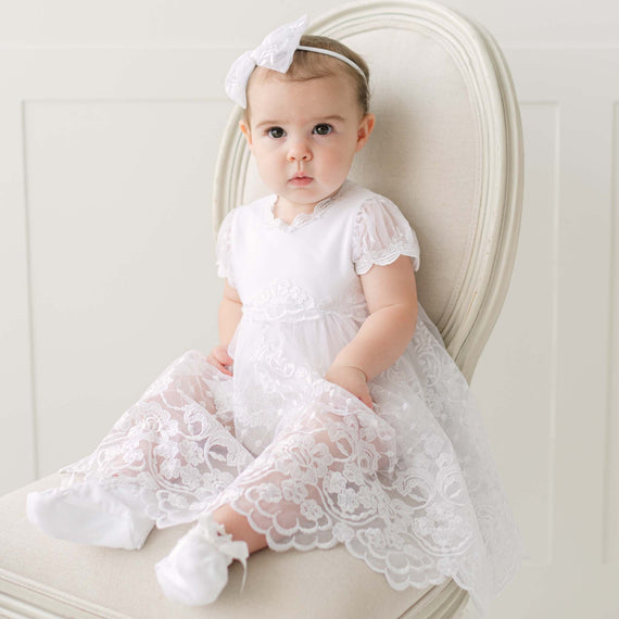 Baby in a white lace dress and a large bow headband sitting on a beige chair.
