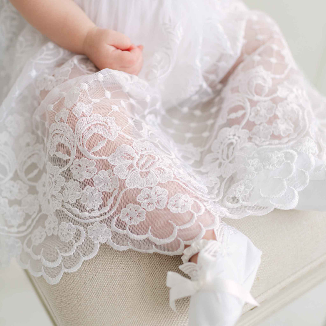 Close-up of a baby's legs in a white lace skirt and booties on a beige background.