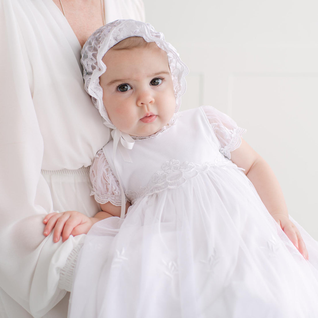 Baby in a white dress and bonnet being held by an adult against a white background.