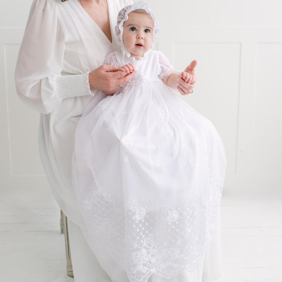 Baby in a white embroidered dress with a bonnet, sitting on her mother's lap.