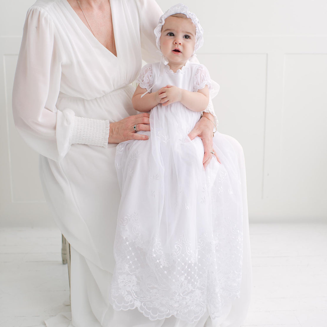 Woman holding a baby dressed in a white gown against a white background.