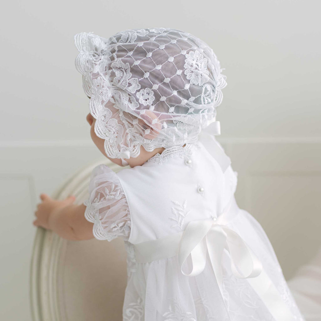Baby wearing a white lace dress and bonnet against a plain background.