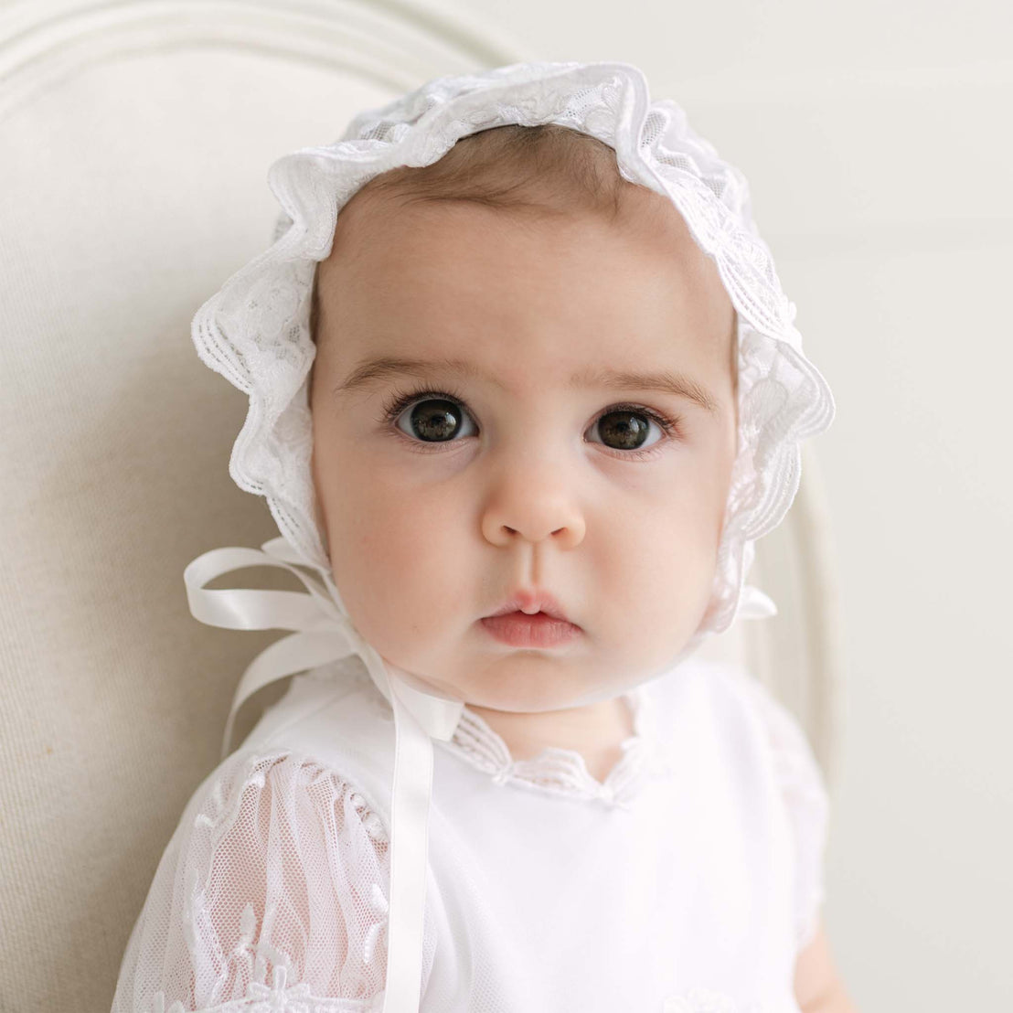 Baby wearing a white lace bonnet against a neutral background.