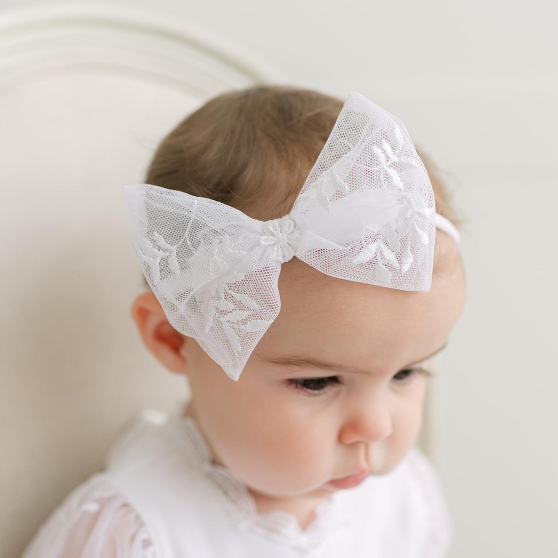 Baby wearing a white lace bow headband against a neutral background.