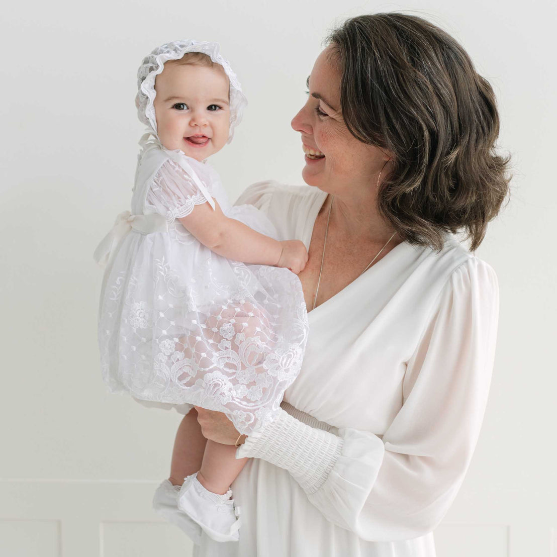 Woman holding a baby dressed in a white dress with a bonnet against a plain background.