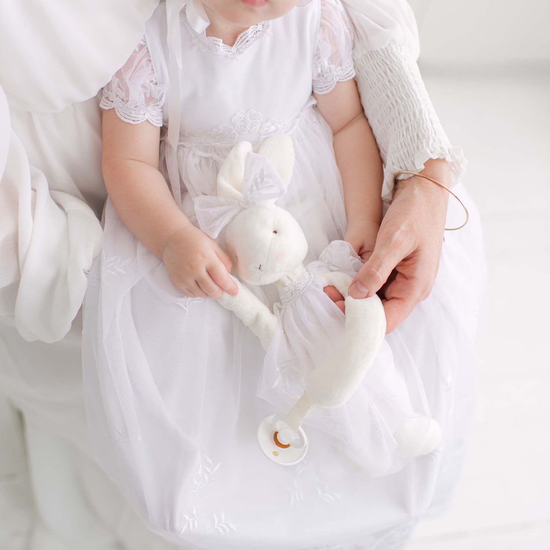Baby in a white dress holding a white plush rabbit against a white background.