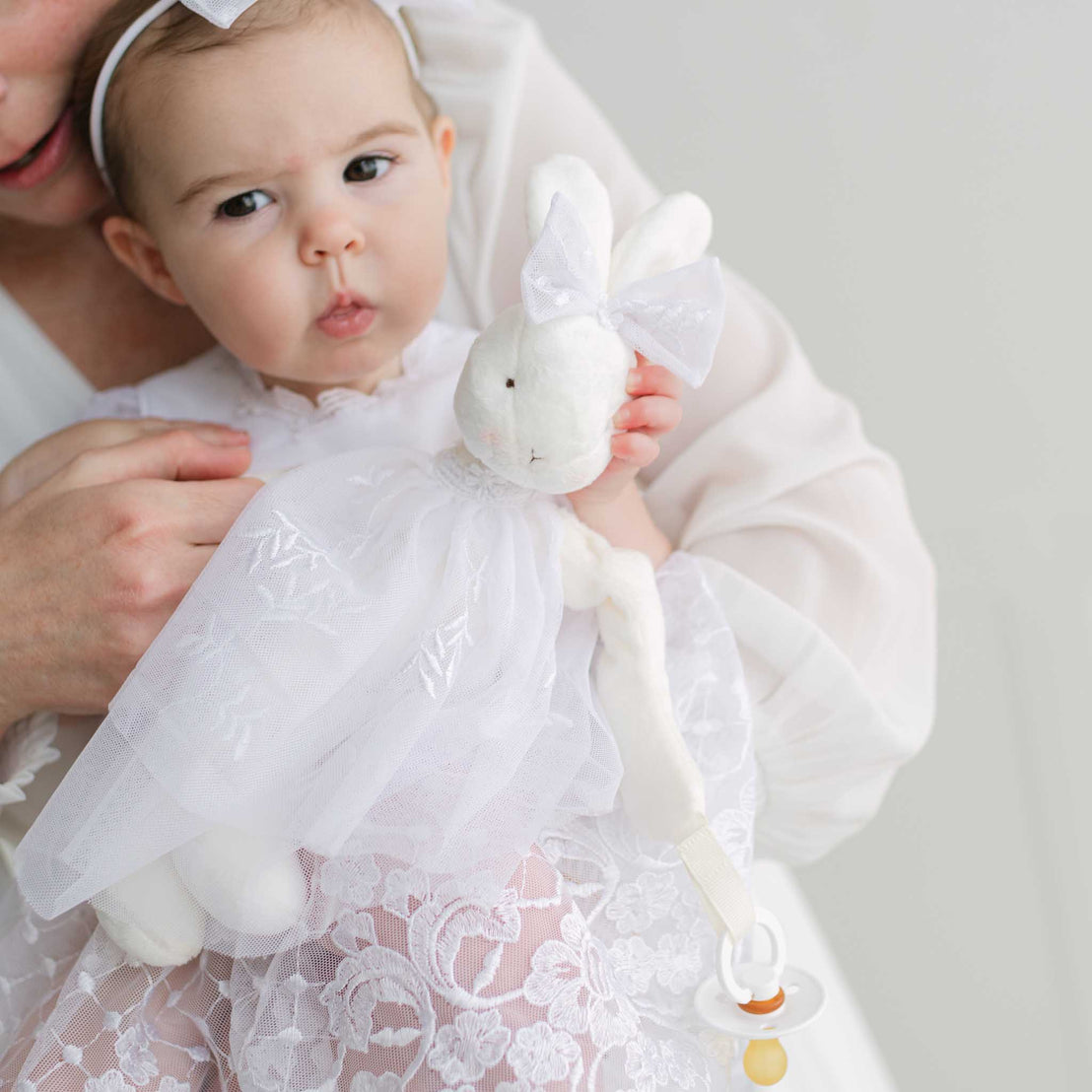 Baby in a white lace dress holding a white rabbit plush toy against a plain background.