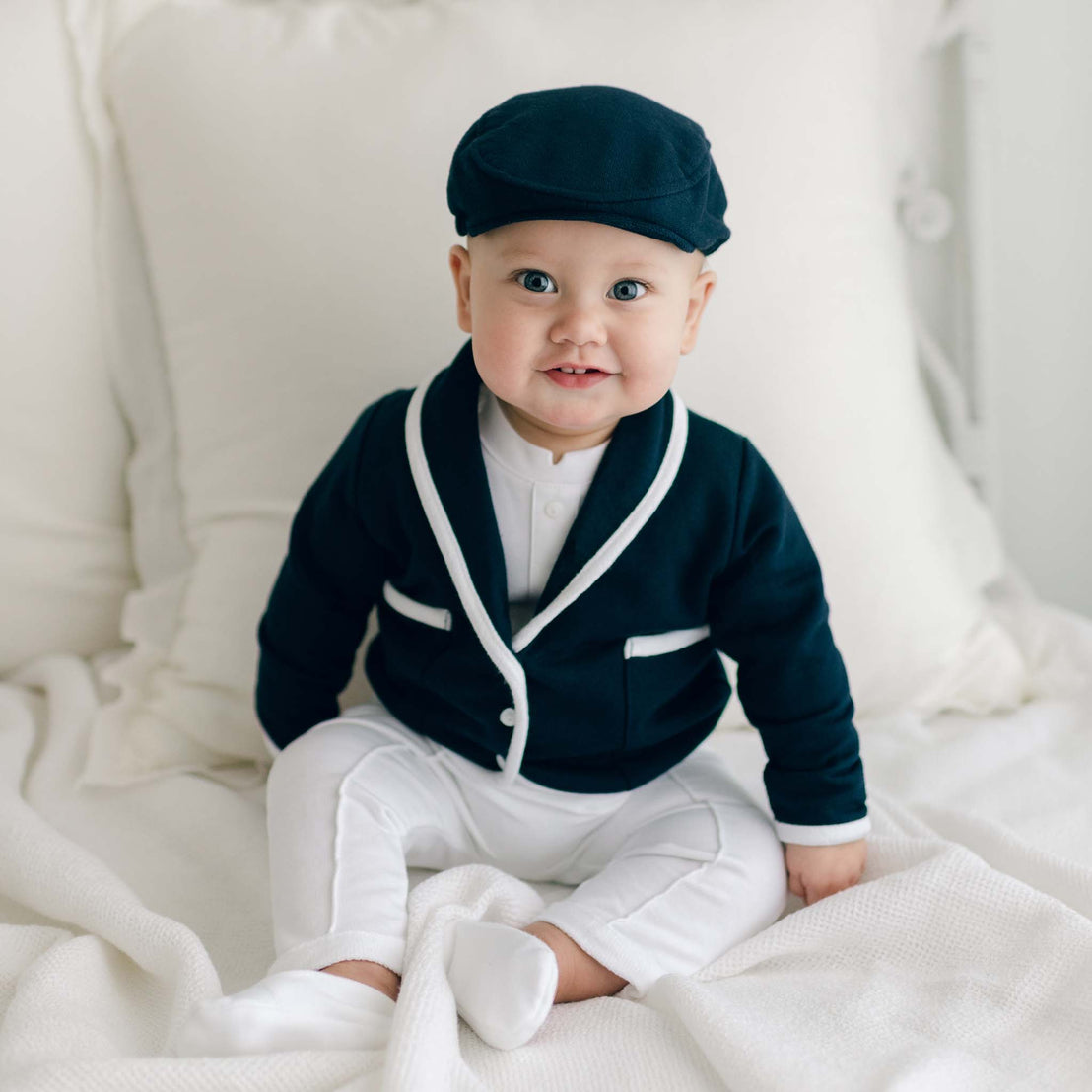 Smiling baby boy sitting on a bed and wearing the Elliott 3-Piece Suit, including the jacket, pants, and onesie (and matching newsboy cap).