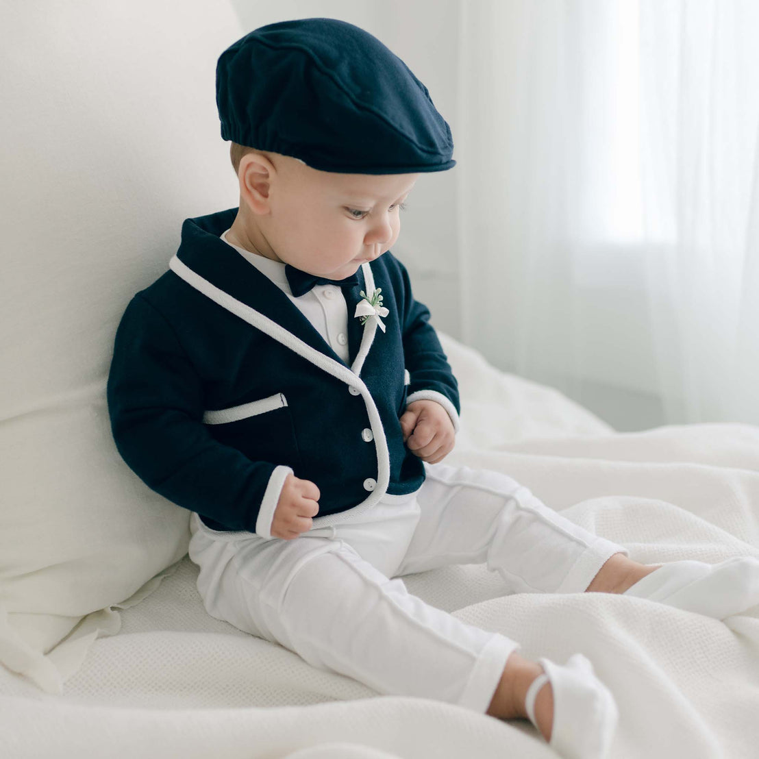 Baby boy sitting on a bed and wearing the Elliott 3-Piece Suit, including the jacket, pants, and onesie (and matching newsboy cap). He is also wearing the Elliott Bow Tie & Boutonniere.