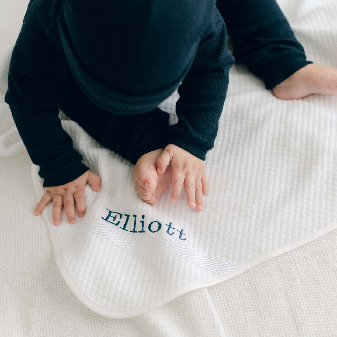 A baby boy in the Navy Ribbed Cotton Top and Legs (including Navy Ribbed Pima Beanie) sitting on the the Elliott Personalized Blanket, a soft white 100% quilted cotton, one corner is detailed with ribbon and the name "Elliott" embroidered in navy.