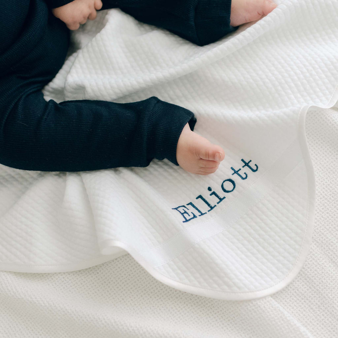 A baby boy sitting on the the Elliott Personalized Blanket, a soft white 100% quilted cotton, one corner is detailed with ribbon and the name "Elliott" embroidered in navy.