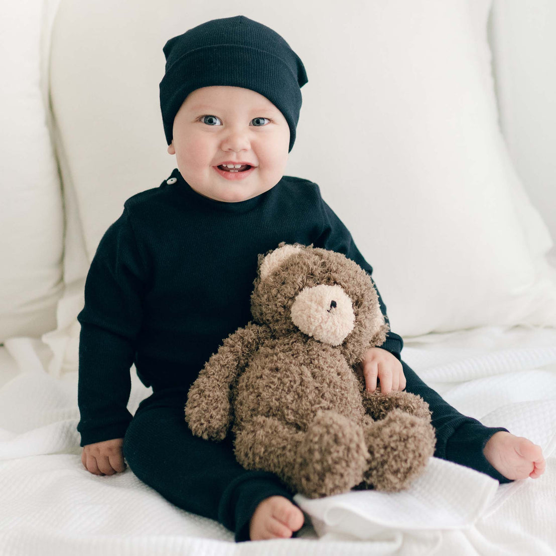 Smiling baby boy sitting the Elliott Personalized Blanket. He is wearing the Elliott Navy Ribbed Cotton Top & Leggings, including the Navy Ribbed Pima Beanie. He is also holding the Cubby Bear.