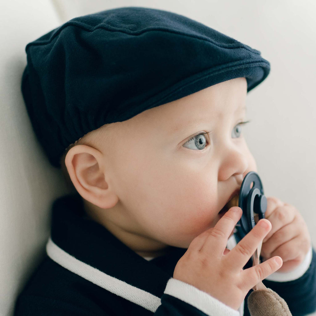 Baby boy with a pacifier in his mouth and wearing the navy Elliott Newsboy Cap.