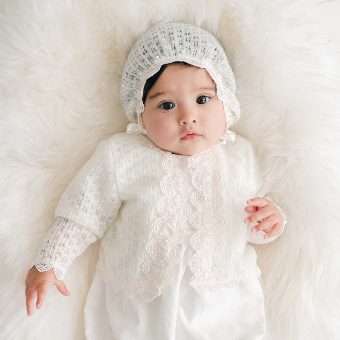 Baby wearing a white knitted outfit and bonnet on a soft white background.