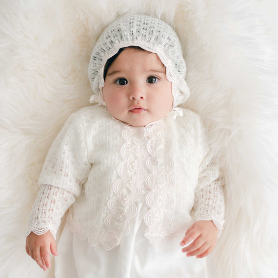 Baby wearing a white knit sweater and bonnet on a soft white background.
