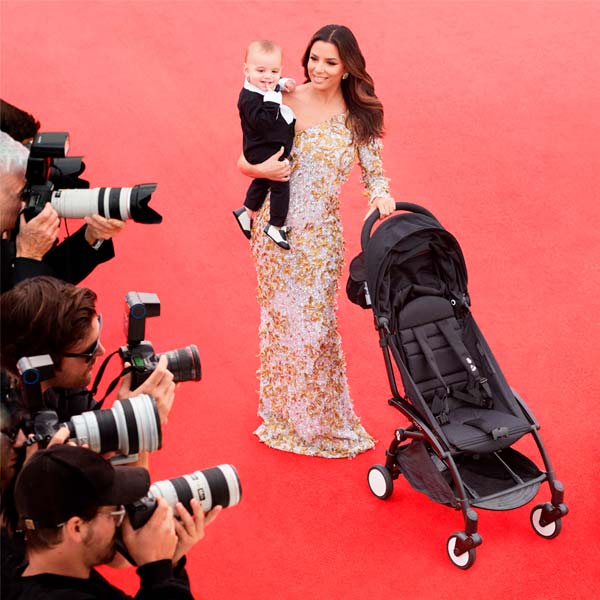 A woman in a glamorous gown holding a child on a red carpet, surrounded by photographers. She is smiling and standing next to a folded stroller.