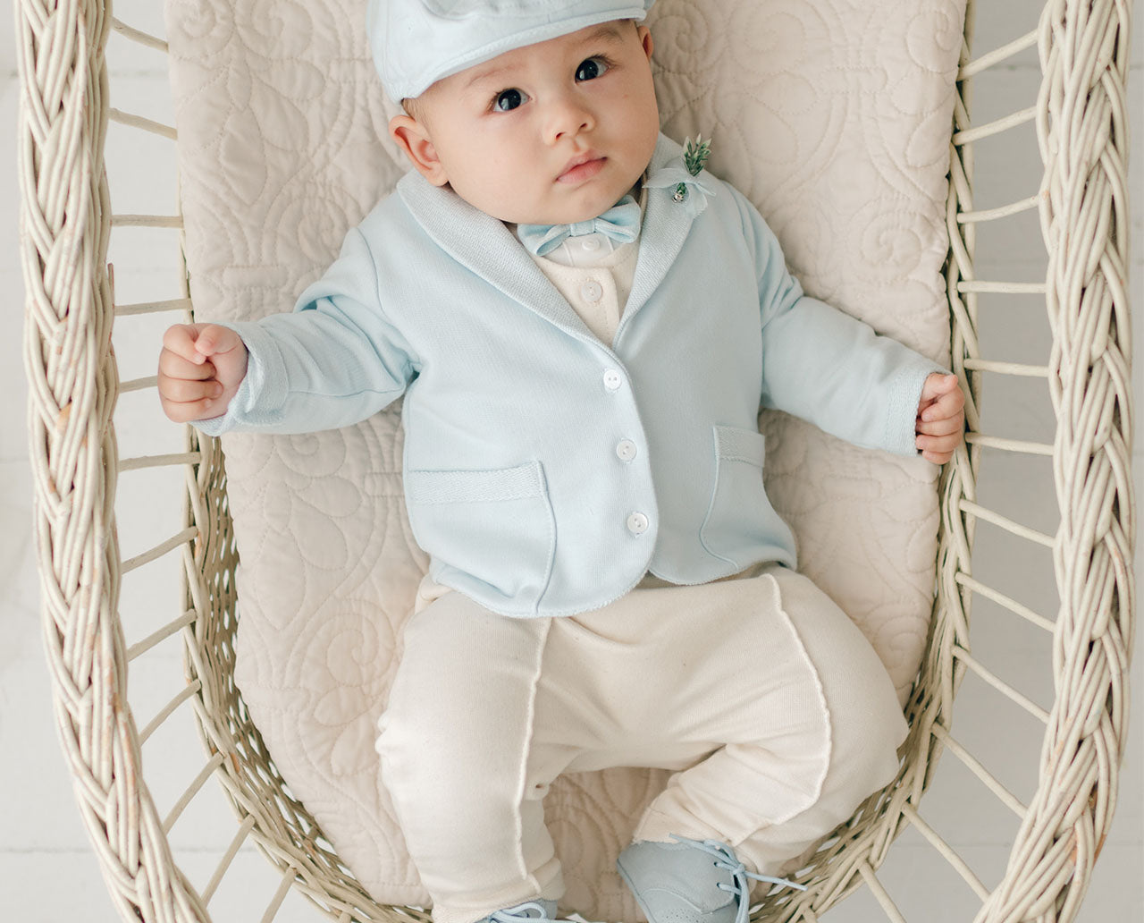 A baby wearing a light blue jacket, cream-colored pants, and a matching light blue hat sits in a woven basket chair. The baby has a small bow tie and looks up with a calm expression, surrounded by soft textures.