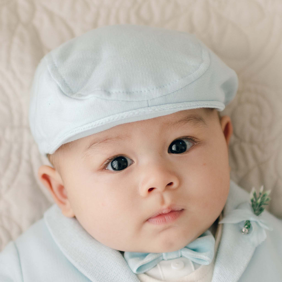 A close-up of a baby wearing an Ezra Powder Blue Newsboy Cap and matching outfit, including a tiny bow tie. The baby has wide, curious eyes and is set against a soft, quilted background, giving off an adorable Christening look.