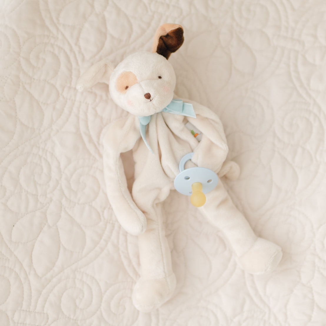 A soft, plush Ezra Powder Blue Silly Puppy Buddy | Pacifier Holder with floppy ears and a pastel blue ribbon around its neck lies on a quilted white blanket. Perfect as a Christening gift, the toy is holding a blue and yellow pacifier.