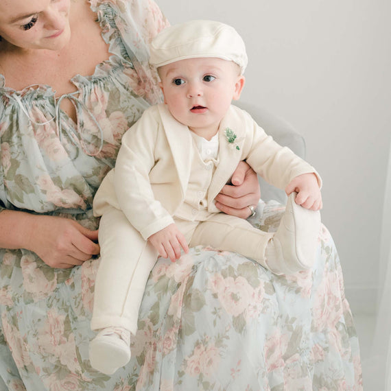 Baby boy sitting on his mother's lap. He is wearing the Ezra 4-Piece Beige Suit with matching Newsboy Cap, and boutonniere