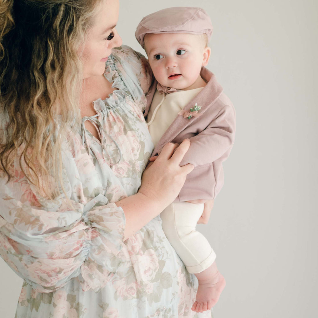 Mother holding her baby boy. The boy is wearing Baby boy standing his mother's lap. He is wearing the Ezra 4-Piece Mauve Suit with matching Newsboy Cap, mauve velvet bow tie and boutonniere 
