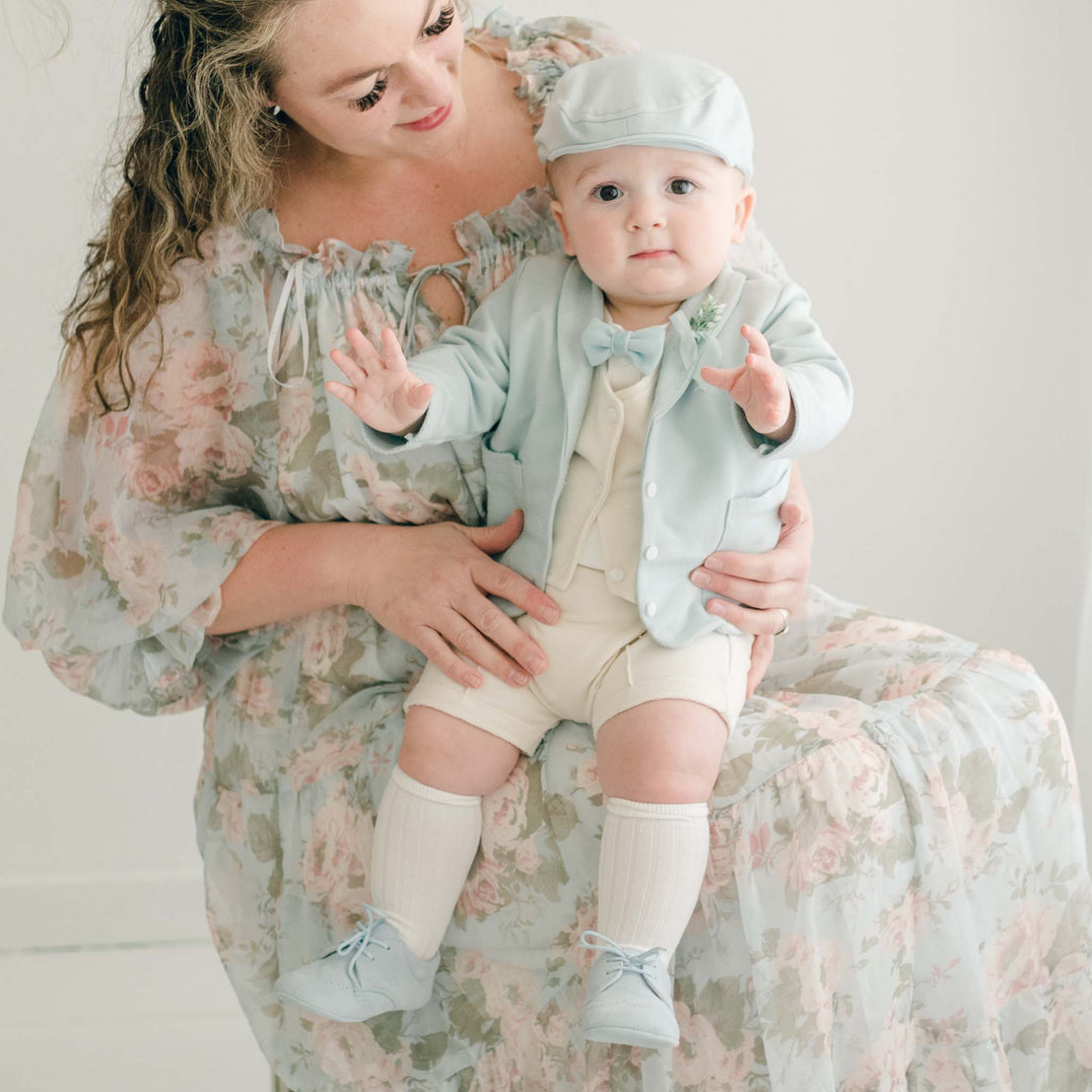 Baby boy sitting on his mother's lap. He is wearing the Ezra 4-Piece Powder Blue Suit with matching Newsboy Cap, blue velvet bow tie and boutonniere