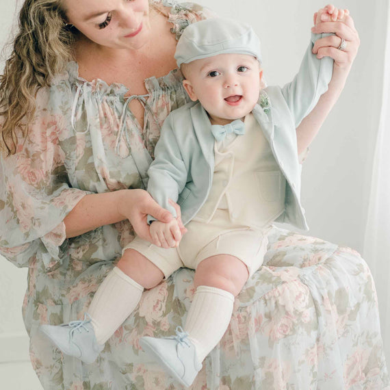 Baby boy sitting on his mother's lap. He is wearing the Ezra 4-Piece Powder Blue Suit with matching Newsboy Cap, blue velvet bow tie and boutonniere