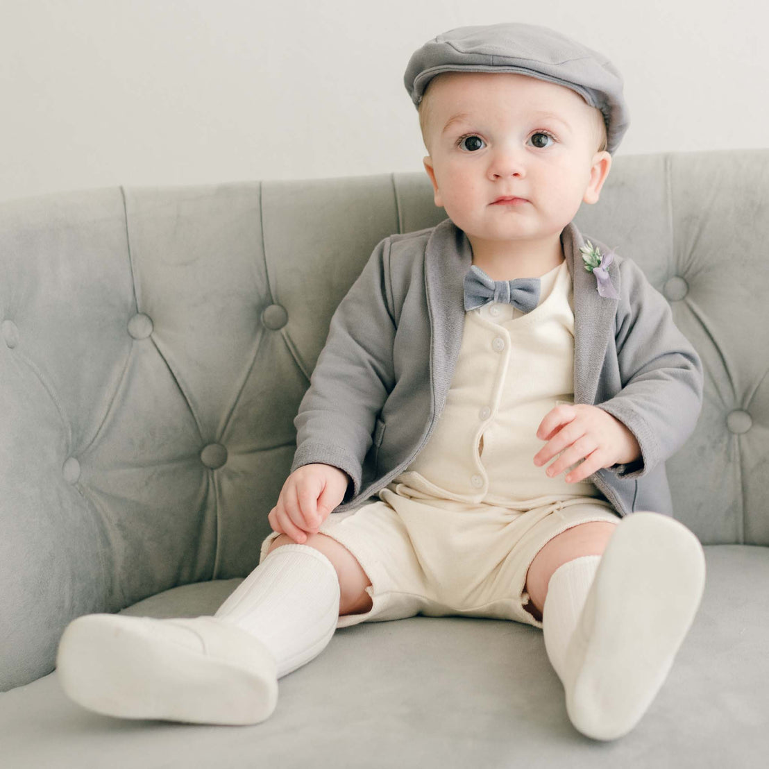 Baby boy sitting on a couch wearing the Ezra 4-Piece Suit with matching Newsboy Cap, heather velvet bow tie and boutonniere