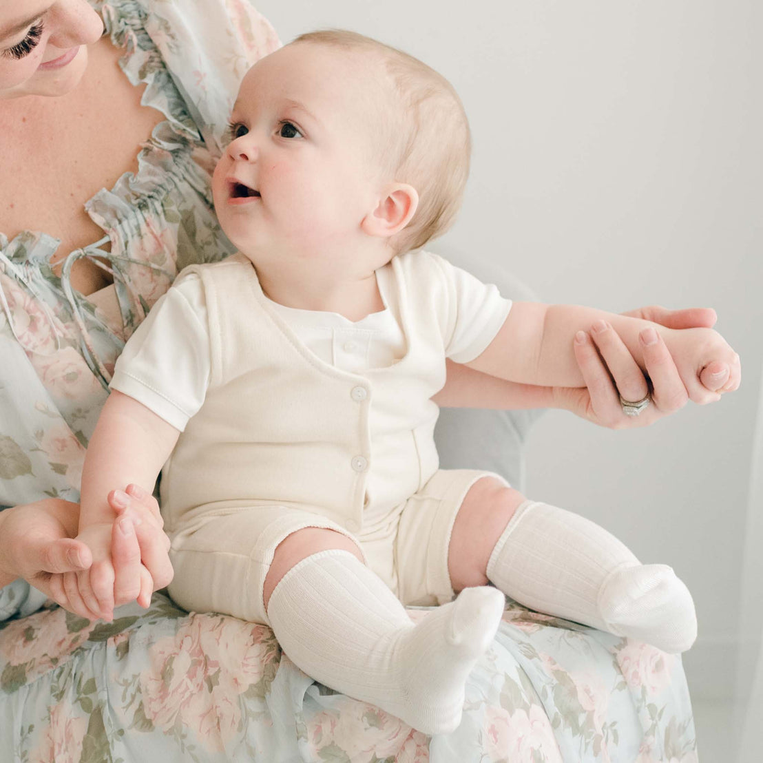 Baby boy sits on his mother's lap. He is wearing a tan vest, shorts and an ivory onesie