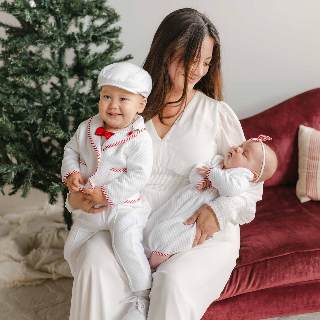 A woman dressed in a white dress sits on a red sofa, holding a baby and a toddler. Both children sport matching Christmas outfits made from quilted cotton; the toddler is wearing the Nicholas 3-Piece Suit, which features red stripes and includes a cap. A beautifully decorated Christmas tree nearby enhances the festive atmosphere.