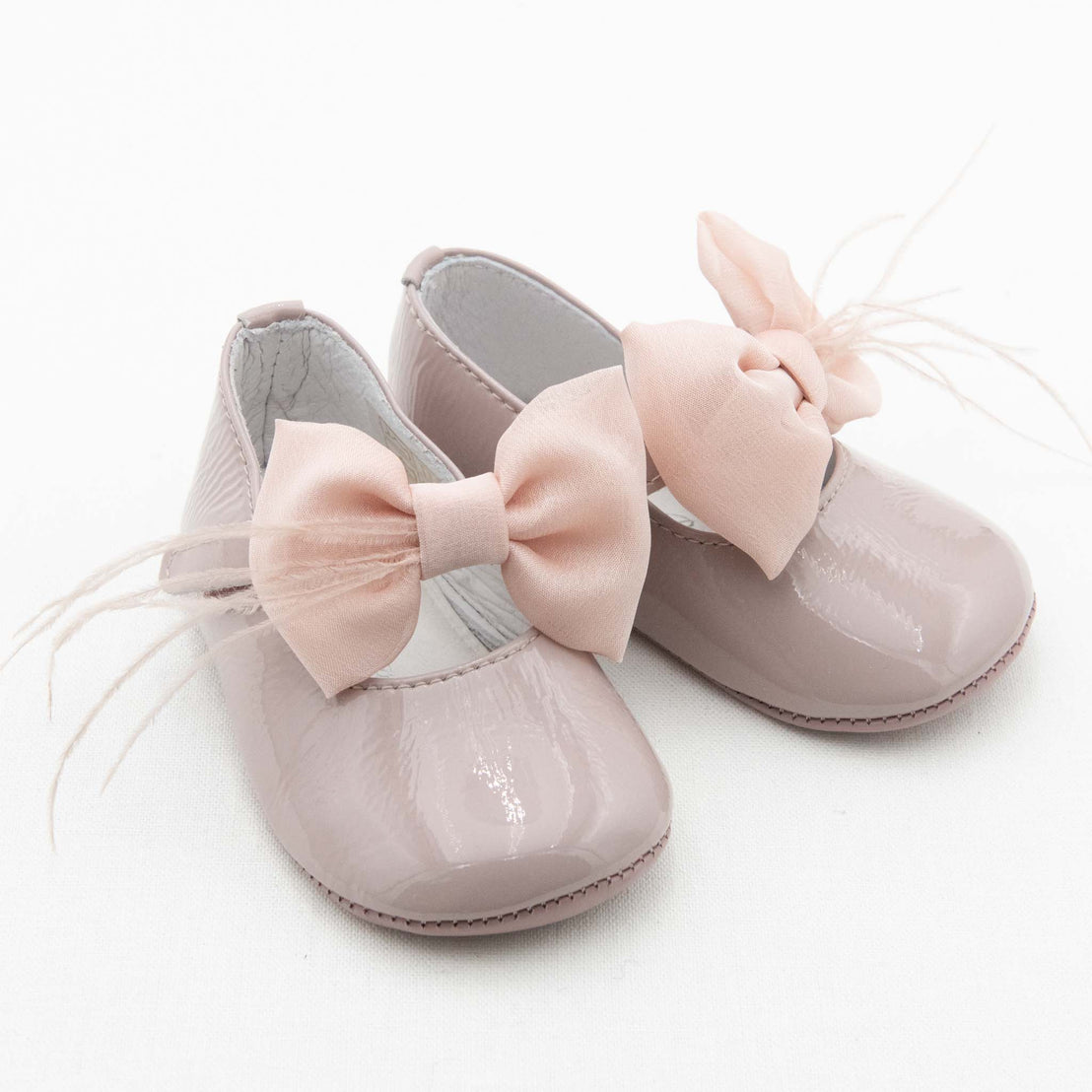 A pair of Rose Patent Leather Mary Janes. The shoes are made with pink patent leather with a soft velcro strap that features a coordinating rose pink chiffon bow and feather. The background is white.