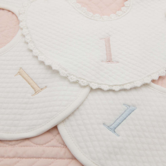 A close-up shot of three handmade, quilted First Birthday Bibs. Each bib is embroidered with the number "1" in pink, blue, or beige. The bibs are arranged overlapping on a pink quilted background. The first bib on the top of the image features a lace trim along its edge.